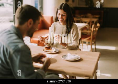 In un caffè caldo e invitante, una coppia condivide risate e deliziose prelibatezze, assaporando caffè e pasticcini mentre si crogiola al sole del pomeriggio. Foto Stock