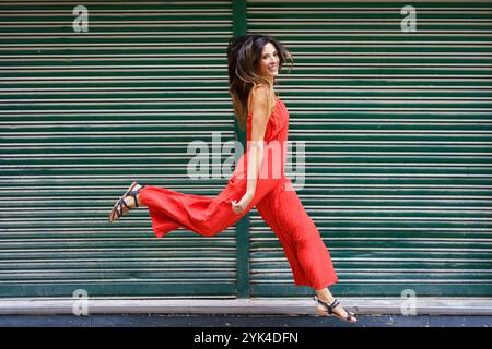Una donna energica vestita con un sorprendente abito rosso è felice di camminare all'aperto in città Foto Stock