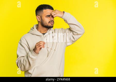 Stanco, stanco, giovane indiano prende gli occhiali, sente dolore agli occhi, essere sovraccarico di lavoro bruciato da lunghe ore di lavoro. Un ragazzo arabo assonnato e stanco che sfrega gli occhi isolato su sfondo giallo Foto Stock
