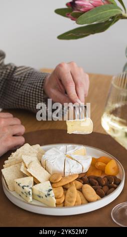 Un bicchiere di vino bianco in mano sullo sfondo di un piatto di formaggi con spuntini e noci Foto Stock