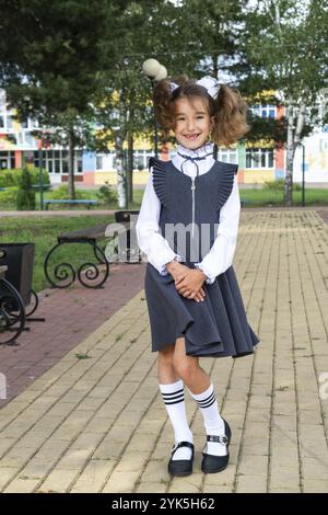 Ragazza allegra e divertente con un sorriso senza denti in uniforme scolastica con archi bianchi nel cortile della scuola. Ritorno a scuola, il 1° settembre. Un allievo felice. Ed. Primaria Foto Stock