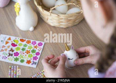 Una ragazza carina con le orecchie rosa coniglietto crea un artigianato pasquale, decora un uovo a forma di unicorno con strass, corno, fiori all'interno di un Foto Stock