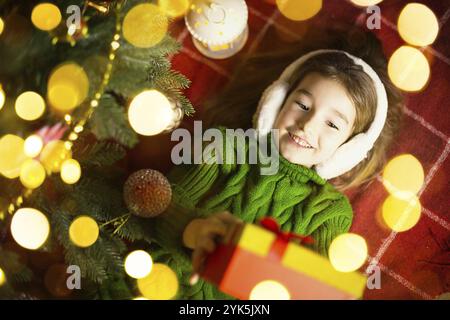 Ragazza in un caldo maglione lavorato a maglia e cuffie in pelliccia è sdraiata su una coperta vicino all'albero di Natale con una scatola regalo. La vigilia di Natale, un sogno e un desiderio. Ne Foto Stock