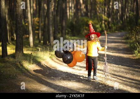 Una ragazza in costume da strega e cappello su una scopa con palloncini arancioni e neri sta giocando nella foresta autunnale, andando ad una festa di Halloween Foto Stock