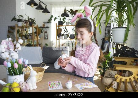 Una ragazza carina con le orecchie rosa coniglietto crea un artigianato pasquale, decora un uovo a forma di unicorno con strass, corno, fiori all'interno di un Foto Stock