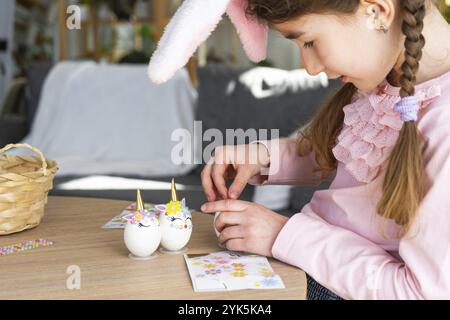 Una ragazza carina con le orecchie rosa coniglietto crea un artigianato pasquale, decora un uovo a forma di unicorno con strass, corno, fiori all'interno di un Foto Stock