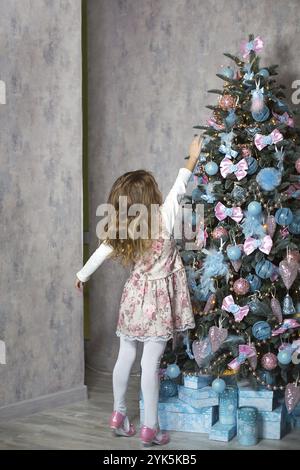 Una bambina raggiunge per un giocattolo dell'albero di Natale con la sua mano. Decorazioni natalizie, in attesa di una vacanza e di un miracolo. Anno nuovo. Bianco, rosa e blu dec Foto Stock