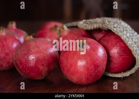 Melograni rossi giacciono su un tavolo di legno. Le mele sono in una borsa intrecciata. Lo sfondo è sfocato con sedie di legno Foto Stock