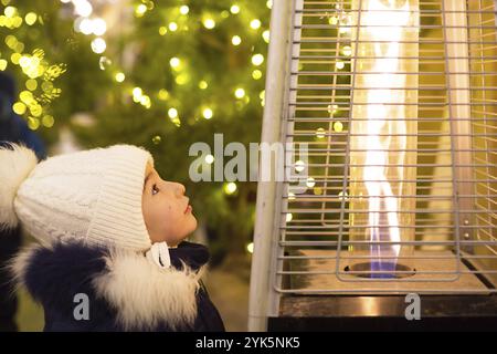 Una bambina in un cappello caldo guarda la fiamma di un bruciatore a gas che si riscalda in un mercatino di Natale in città. luci di ghirlande in sfocatura sul christm Foto Stock