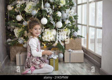 Bambina in bel vestito è seduta sotto l'albero di Natale con scatola regalo e arco. Luce da una grande finestra, camera buia, comfort di casa, Natale, m Foto Stock