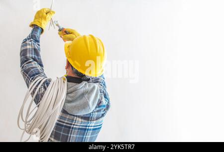 Un elettricista con elmetto giallo e guanti lavora sull'installazione del cablaggio elettrico, dimostrando le pratiche di sicurezza in un ambiente ben illuminato. Foto Stock