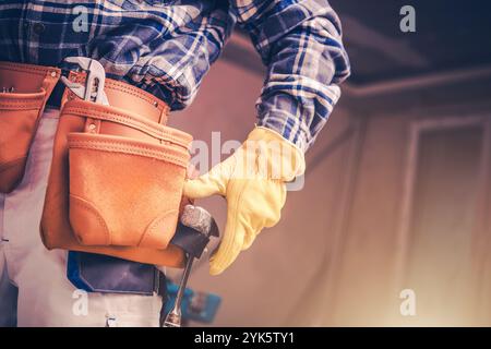 Un lavoratore esperto, vestito con una camicia a quadri e guanti, recupera un martello da una cintura portautensili, pronto per un'attività di costruzione in un ambiente interno ben illuminato Foto Stock