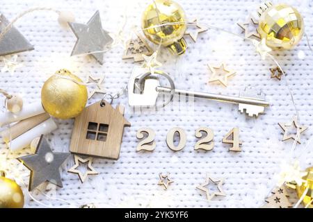 Chiave di casa con cottage portachiavi su sfondo festivo con stelle e luci di ghirlande. Capodanno 2024 lettere di legno, biglietto d'auguri. Acquisto, costruzione Foto Stock