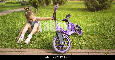 Ragazza allegra seduta sull'erba in un parco vicino alla bici da corsa viola nel periodo estivo. Animazione per bambini, scooter per todd Foto Stock