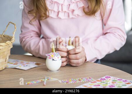 Una ragazza carina con le orecchie rosa coniglietto crea un artigianato pasquale, decora un uovo a forma di unicorno con strass, corno, fiori all'interno di un Foto Stock