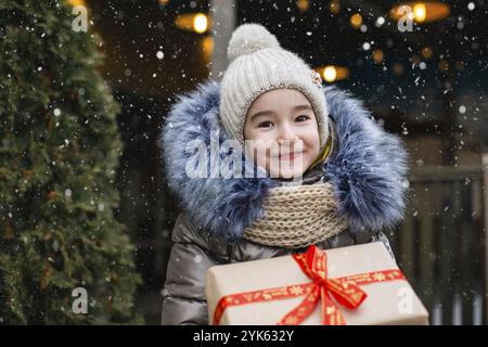 Ritratto di ragazza gioiosa con una scatola regalo per Natale su una strada cittadina in inverno con neve su un mercato festivo con decorazioni e luci. Panno caldo Foto Stock