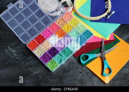 Progetto fai-da-te in fase di realizzazione. Foto dall'alto di perline di vetro colorate, tessuto in feltro e forbici sulla scrivania grigio scuro. Lavoro manuale in corso. Foto Stock