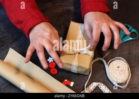 Mani di una ragazza in un abito rosso che decora una confezione regalo con nastro in pizzo. Mani femminili che tengono in mano una confezione regalo. Foto Stock