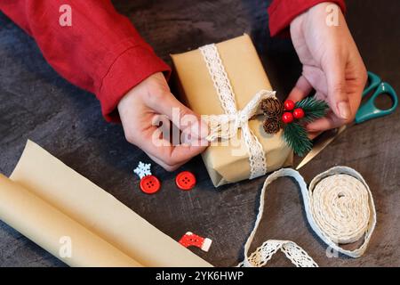 Mani di una ragazza in un abito rosso che decora una confezione regalo con nastro in pizzo. Mani femminili che tengono in mano una confezione regalo. Foto Stock