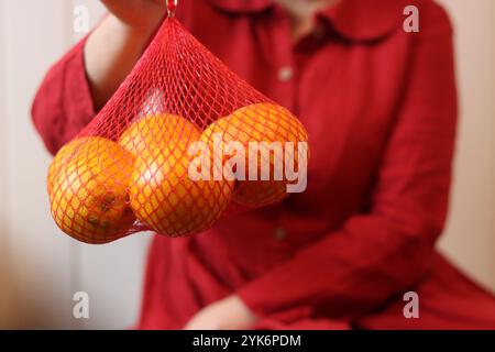 Donna in abito rosso con arance in borsa mesh su sfondo chiaro, primo piano delle mani Foto Stock