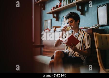 Giovane che si gode un drink mentre legge un libro a casa Foto Stock