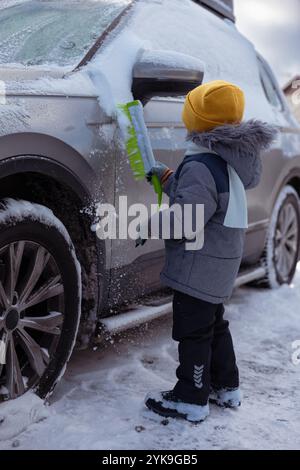 Bambino con abiti invernali caldi che puliscono la neve dall'auto di famiglia con una spazzola in una giornata fredda e gelida. Bambino che aiuta i genitori e la manutenzione invernale Foto Stock