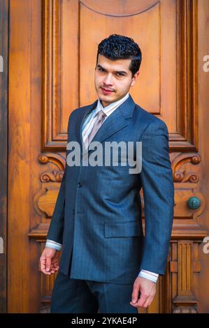 Un uomo con una tuta di pinstrime si erge con sicurezza davanti a una porta di legno riccamente intagliata. Ha un leggero sorriso e indossa una cravatta di colore chiaro, ADD Foto Stock