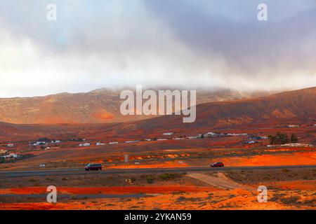 Terreno arido di Fuerteventura, strada con pochi veicoli che vi viaggiano. Montagne sullo sfondo, ci sono basse colline parzialmente coperte da clou Foto Stock