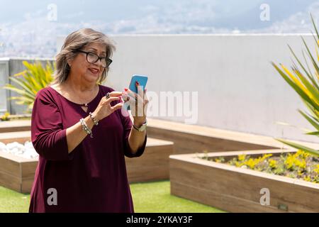 Una donna latina anziana che guarda il suo cellulare sorridendo in giardino la mattina Foto Stock