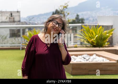 Donna anziana in camicetta Bordeaux che parla al telefono cellulare sulla terrazza in una mattina d'estate Foto Stock