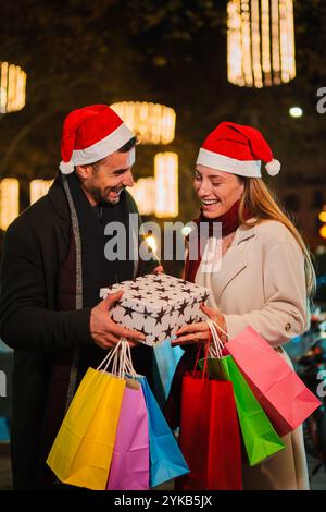 Verticale. Fidanzato che fa un regalo di Natale alla sua ragazza all'aperto di notte, indossando cappelli di Babbo Natale e sorridendo di gioia, circondato da luci festive e borse dello shopping durante le festività natalizie. Foto di alta qualità Foto Stock