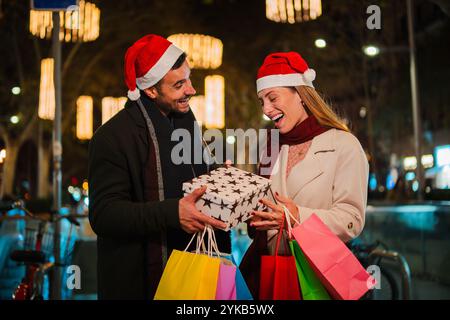 Fidanzato che fa un regalo di Natale alla sua ragazza all'aperto di notte, indossando cappelli di Babbo Natale e sorridendo di gioia, circondato da luci festive e borse dello shopping durante le festività natalizie. Foto di alta qualità Foto Stock