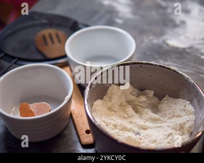 Farina, uova e utensili sparsi su un piano di lavoro spolverato di farina suggeriscono di preparare biscotti in corso. Questa accogliente scena cattura tutto l'essenziale per preparare biscotti Foto Stock