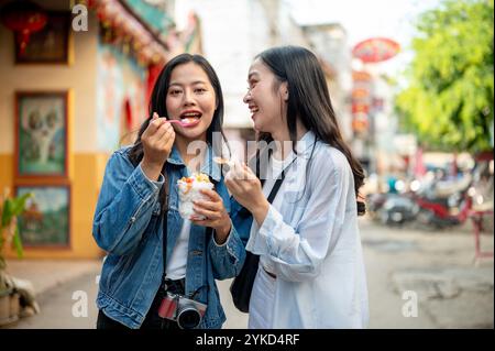 Due graziose e gioiose giovani turisti asiatiche si godono il cibo di strada in un mercato in Thailandia, mangiando gelato al cocco insieme per strada durante il loro soggiorno Foto Stock