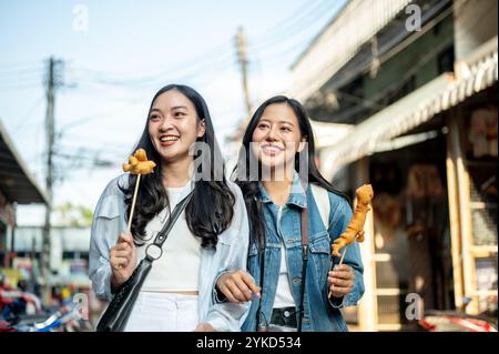 Due gioiose turisti asiatici si godono una passeggiata in città in una giornata di sole, passeggiando lungo la strada in un mercato in Thailandia e mangiando cibo di strada. S Foto Stock