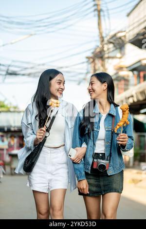 Due gioiose turisti asiatici si godono una passeggiata in città in una giornata di sole, passeggiando lungo la strada in un mercato in Thailandia e mangiando cibo di strada. S Foto Stock