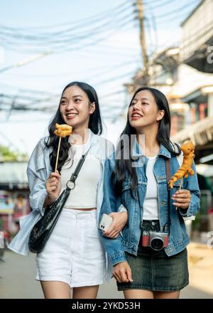 Due gioiose turisti asiatici si godono una passeggiata in città in una giornata di sole, passeggiando lungo la strada in un mercato in Thailandia e mangiando cibo di strada. S Foto Stock