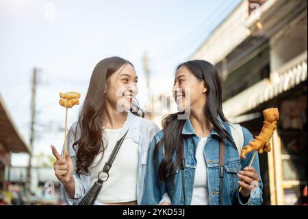 Due gioiose turisti asiatici si godono una passeggiata in città in una giornata di sole, passeggiando lungo la strada in un mercato in Thailandia e mangiando cibo di strada. S Foto Stock