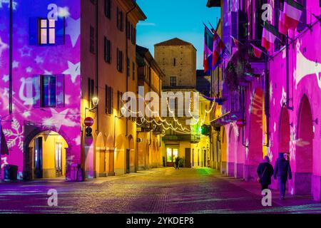 Piccola città italiana di Alba al tramonto, con una stretta strada acciottolata, edifici storici illuminati da calde luci natalizie la sera. Foto Stock