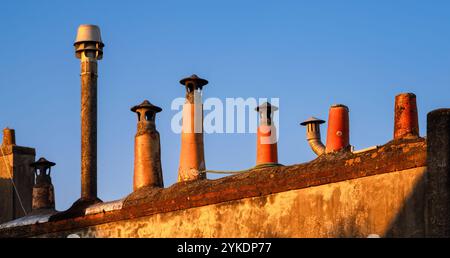 Una fila di vari camini si staglia su un tetto contro un cielo azzurro. I camini variano in altezza e design. La calda luce del tramonto proietta un dorato Foto Stock