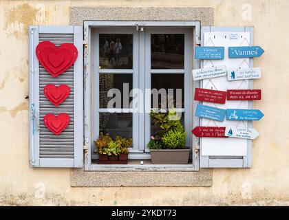 Vannes, Francia - 27 luglio 2024: Un'affascinante finestra con persiane blu adornata con decorazioni a cuore rosso e vari segni colorati. Le piante in vaso si siedono Foto Stock