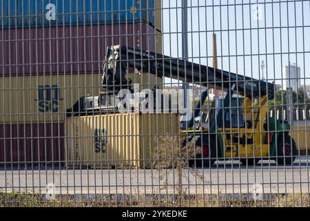 Raggiungere il raccoglitore sollevando un contenitore di spedizione dietro una recinzione a rete metallica in una porta Foto Stock