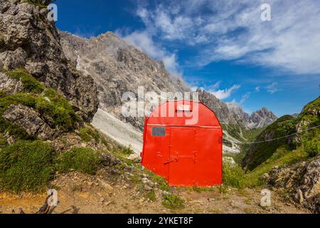 Rifugio alpino bivaco situato in un aspro terreno alpino. Rifugio rosso bivacco nelle Dolomiti. Struttura compatta contro paesaggi mozzafiato Foto Stock