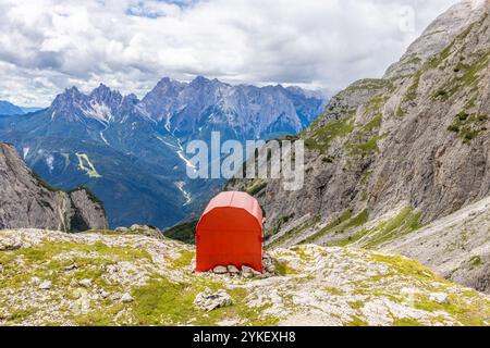 Rifugio alpino bivaco situato in un aspro terreno alpino. Rifugio rosso bivacco nelle Dolomiti. Struttura compatta contro paesaggi mozzafiato Foto Stock