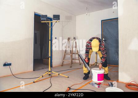 Un lavoratore mescola la vernice in un secchio mentre si prepara a dipingere le pareti in una stanza in costruzione. Gli strumenti e le luci sono disposti intorno allo spazio. Foto Stock