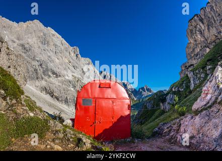 Rifugio alpino bivaco situato in un aspro terreno alpino. Rifugio rosso bivacco nelle Dolomiti. Struttura compatta contro paesaggi mozzafiato Foto Stock