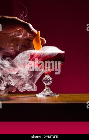 Un elegante cocktail Negroni servito in un bicchiere di cristallo, avvolto da fumo aromatico, Una mano aggiunge un tocco di agrumi, esaltando le vivaci sfumature di rosso Foto Stock