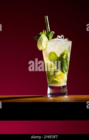 Un vivace cocktail mojito guarnito con menta fresca e una fetta di lime in un bicchiere alto. La bevanda è collocata su un ricco sfondo rosso, evidenziandone la presenza Foto Stock