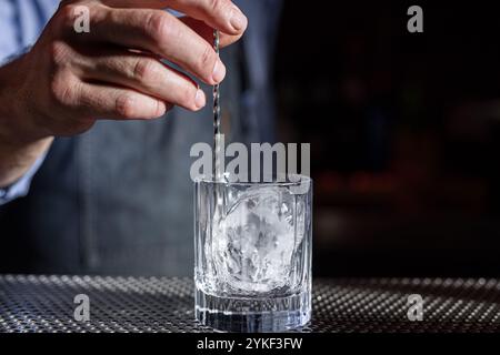 Un barista inizia preparando un classico whisky vecchio stile con un cucchiaio di metallo sul ghiaccio, il vetro trasparente e lo sfondo scuro mettono in risalto l'eleganza Foto Stock