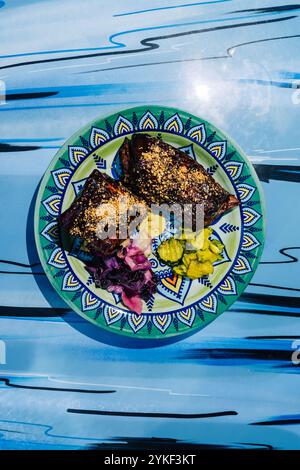 vista dall'alto di succulenti spareribs placcati con sottaceti, sciamano di cavolo e salse su un piatto in ceramica dal design vivace, adagiato su un intrigante bac blu Foto Stock